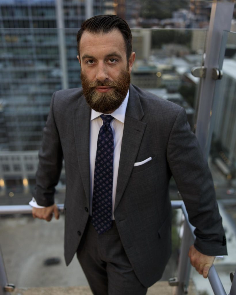 Michael J. Penney wearing a grey suit and blue tie with pink accents, a white shirt, standing with the city behind him
